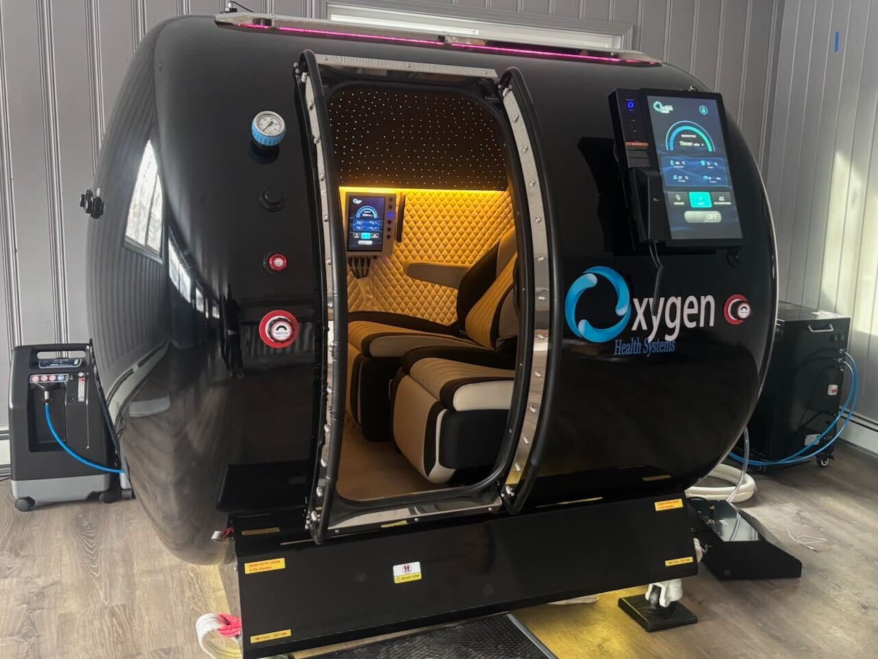 A supportive technician assists a relaxed client inside a transparent hyperbaric oxygen chamber in a bright, serene wellness center room with digital controls, medical equipment, and clean white tile floor, illustrating a professional yet calming atmosphere for hyperbaric therapy.